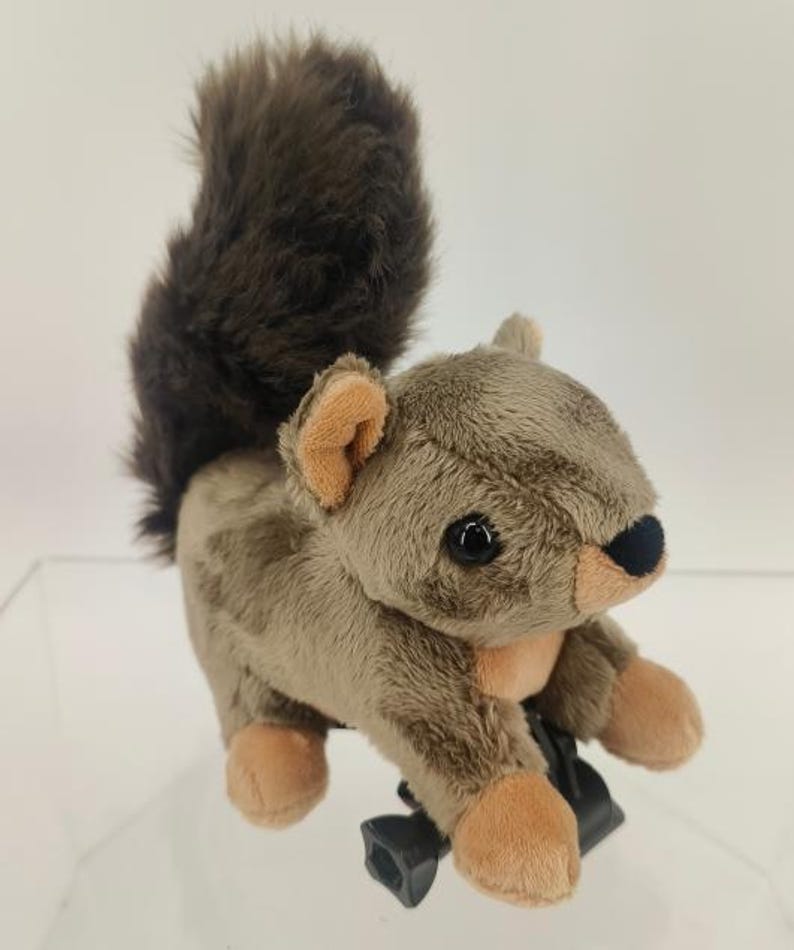 May include: A plush squirrel toy with a fluffy brown tail and tan body. The toy has a black nose, black eyes, and tan paws and ears. The squirrel is attached to a black plastic clip.