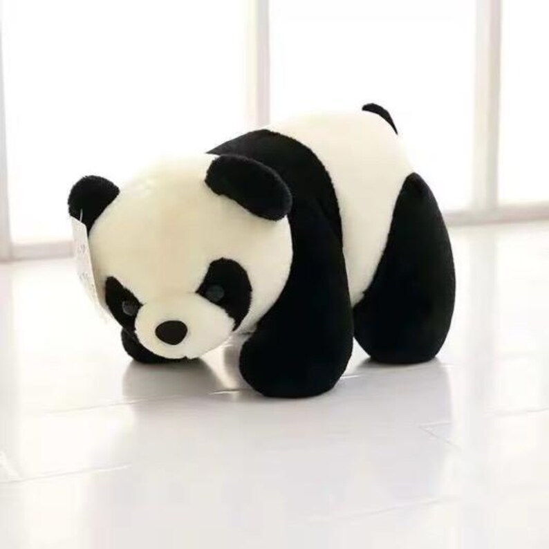 May include: A plush panda bear toy in a playful pose. The panda has a black and white color scheme, with black ears, eye patches, and limbs. The soft toy is made of a fluffy material and is positioned on a white surface.