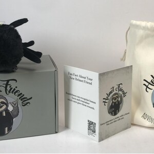 May include: A plush bumblebee toy with black, yellow, and white accents sits atop a gray box. The box and a drawstring bag feature the "Helmet Friends" logo with a sloth illustration. A folded card with the same logo is also present.