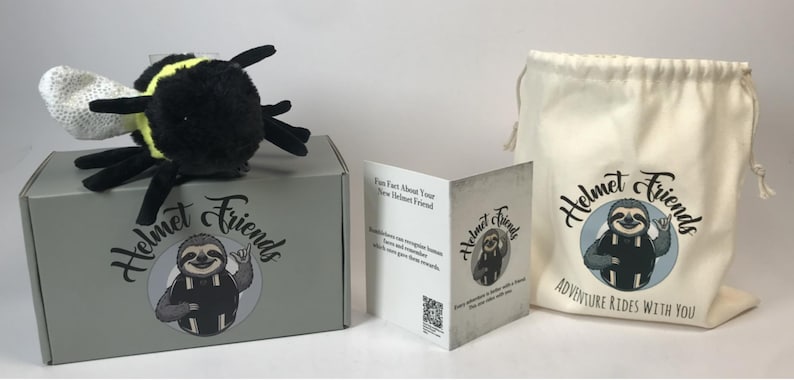 May include: A plush bumblebee toy with black, yellow, and white accents sits atop a gray box. The box and a drawstring bag feature the "Helmet Friends" logo with a sloth illustration. A folded card with the same logo is also present.