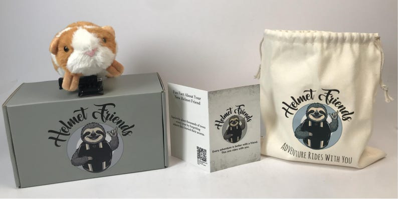 May include: A plush guinea pig helmet accessory sits atop a gray box with the "Helmet Friends" logo. A drawstring bag and a folded card with the same logo are also visible. The bag reads "Adventure Rides With You."