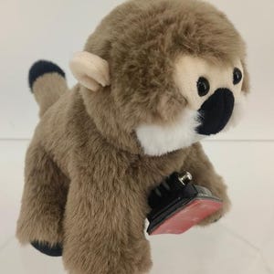 May include: A plush monkey toy with brown fur, a white face, and black eyes and nose. The monkey has a black and blue tail and is attached to a black and red base.