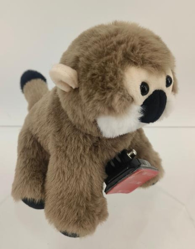 May include: A plush monkey toy with brown fur, a white face, and black eyes and nose. The monkey has a black and blue tail and is attached to a black and red base.