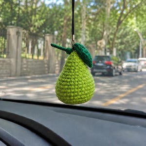May include: A lime green crocheted pear ornament hanging from a black cord. The pear features a textured, knotted surface with a small, dark green crocheted leaf attached to the top stem. The object is suspended in front of a car windshield.