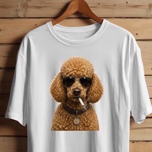 Op de afbeelding: Een wit t-shirt met korte mouwen en een grafische print van een bruine poedel met een zwarte zonnebril en een halsband met een penning, terwijl het een sigaret in de bek houdt. Het t-shirt hangt aan een houten kledinghanger tegen een houten achtergrond.