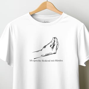 May include: A white short-sleeved t-shirt featuring a black line drawing of a hand making a gesture and the text "Ich spreche fließend mit Händen" printed in black ink on the centre of the chest area.