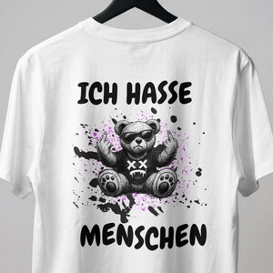 Könnte beinhalten: Ein weißes T-Shirt mit kurzen Ärmeln und einem Aufdruck eines Teddybären, der eine Sonnenbrille trägt und den Mittelfinger zeigt. Der Bär ist von violetten Farbspritzern umgeben. Der Text "ICH HASSE MENSCHEN" ist in Schwarz aufgedruckt.