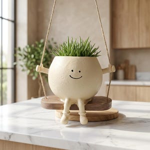May include: A cream-colored ceramic planter shaped like a round character with a simple smiling face, arms, and legs. The planter sits on a small wooden swing suspended by thin ropes, containing a cluster of spiky green succulent-like foliage.