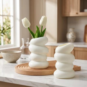 Könnte beinhalten: Zwei weiße Keramikvasen mit einer gestapelten, abgerundeten, spiralförmigen Form. Die größere Vase enthält zwei weiße Tulpen mit grünen Stielen und Blättern. Die Vasen haben eine glatte, matte Oberfläche und eine markante, wellenförmige Silhouette.