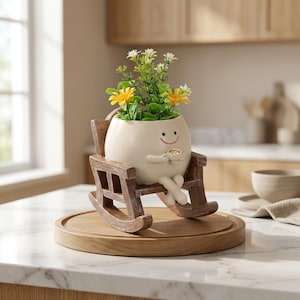 May include: A cream-colored, round planter shaped like a character with a smiling face, holding a small cup, and sitting in a brown wooden rocking chair. The planter contains a cluster of green foliage with small yellow and white flowers.