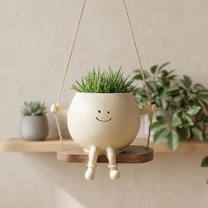 Pode incluir: Um vaso de cerâmica creme com a forma de um personagem sorridente, completo com braços, pernas e um assento de balanço de madeira. O vaso contém plantas verdes semelhantes a grama. O vaso é suspenso por uma corda, criando um elemento decorativo lúdico.