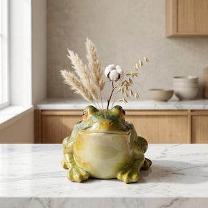 Puede incluir: Una maceta de cerámica con forma de rana y acabado brillante en verde y amarillo. La rana tiene grandes ojos con borde naranja y una boca ancha. Contiene un arreglo decorativo de hierba de la pampa beige, tallos de avena y una flor de algodón.