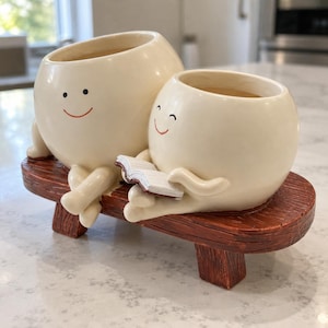 Puede incluir: Dos macetas de cerámica color crema, redondas y sonrientes, se asientan sobre un banco marrón con textura de madera. La maceta más pequeña a la derecha sostiene un pequeño libro blanco abierto con bordes marrones. Ambas tienen rostros pintados.