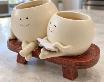 May include: Two cream-colored, round, smiling ceramic planters sit on a brown, wood-textured bench. The smaller planter on the right holds a small, open white book with brown edges. Both planters feature simple, hand-painted facial expressions.