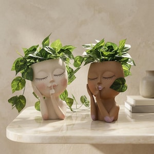 May include: Two head-shaped planters, one in cream and one in brown, each featuring a face with closed eyes and a finger pressed to the lips in a shush gesture. Both planters are filled with green trailing foliage and have pink blush accents.