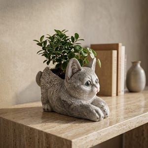 Op de afbeelding: Een grijze, katvormige plantenbak met textuurdetails van vacht, met een kleine groene plant die uit de opening op de rug groeit. De kat is afgebeeld in een liggende positie met de voorpoten naar voren gestrekt en het hoofd licht gedraaid.