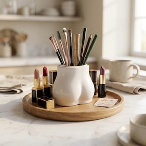 May include: A white ceramic holder shaped like buttocks sits on a round wooden tray, filled with various makeup brushes and pencils. Several lipsticks in black and gold cases are arranged around the holder, alongside a small square card.