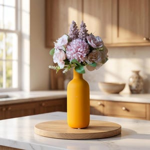 May include: A bright yellow cylindrical vase holds a bouquet of artificial flowers in shades of light pink, mauve, and cream, featuring a central pom-pom bloom, surrounding roses, and two tall, textured, lavender-colored floral spikes.