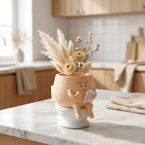 May include: A peach-colored ceramic planter shaped like a character sitting on a white toilet, holding a white tissue. The planter is filled with a bouquet of dried beige grasses and flowers, featuring a textured surface and a small angry facial expression.