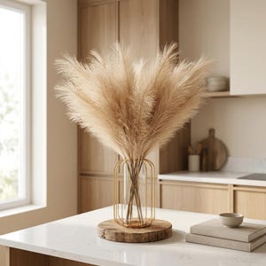 May include: A decorative arrangement of dried pampas grass in a clear glass vase with a gold metal cage design. The vase sits on a round wooden base. The arrangement is on a white countertop in a kitchen setting.