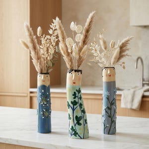 May include: Three tall ceramic vases shaped like human figures, each featuring a painted face at the top and floral or botanical patterns on the body. The vases are filled with arrangements of dried grasses, pampas, and white seed pods.