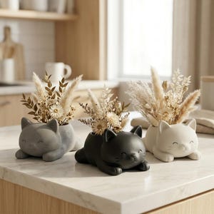 Peut inclure: Trois cache-pots en forme de chat de couleur grise, noire et blanche, chacun présentant une finition lisse et un visage souriant. Ces récipients en céramique sont remplis d'arrangements d'herbes séchées beiges et de petites fleurs blanches.