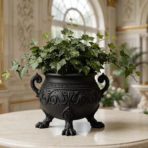 May include: A matte black planter with ornate embossed scrollwork, two side handles, and three clawed feet. The planter contains a lush green ivy plant with trailing vines extending over the edges of the rounded, decorative vessel.