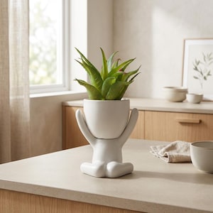 May include: A white ceramic planter in the shape of a person holding a green plant. The planter has a minimalist design with arms and legs. The plant has green leaves. The planter is on a light-colored countertop.