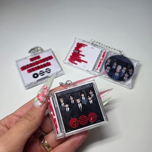 May include: Clear plastic CD-shaped keychains featuring images of a musical group. The keychains have red and black accents, with text that reads "BTS WORLD TOUR" and "ARRANG". The keychains are attached to a silver chain.