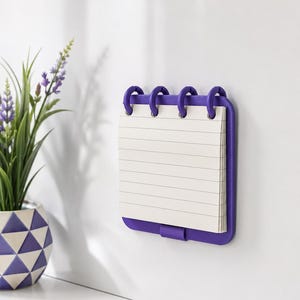Puede incluir: Un bloc de notas magnético cuadrado de color morado con páginas de papel blanco rayado unidas por cuatro anillas de plástico moradas. El bloc tiene un soporte de plástico morado sólido y un pequeño clip morado en el centro inferior.