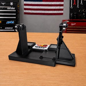 May include: A black metal wheel truing stand featuring two vertical supports with adjustable rollers at the top. A white rectangular plate is centered on the base, displaying the text "SKORTS RACING" in black with a red and yellow flame graphic.