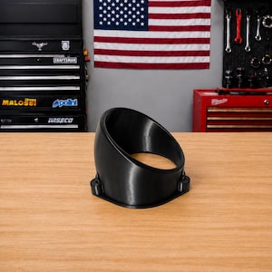 May include: A black, cylindrical gauge pod housing with an angled, circular opening at the top and a flat, square-shaped base featuring two small mounting holes on opposite sides, constructed from a smooth, solid material with a matte finish.