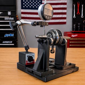 May include: A black metal crankshaft truing stand featuring a digital dial indicator mounted on an adjustable arm. The base includes a magnetic switch labeled with the text SKOOTS and a flame graphic. The device holds a steel crankshaft assembly.