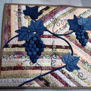 May include: A quilted wall hanging featuring a grape vine design. The grapes are a dark blue, with matching leaves and stems. The background is made of various fabric strips in shades of brown, pink, and cream, with a brown border.