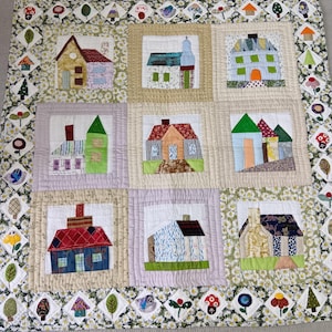Op de afbeelding: Een vierkante patchwork quilt met negen centrale blokken, elk met een ander huisontwerp in diverse stoffen en kleuren. De quilt is omlijst door een rand van kleine ruitvormige lapjes met afbeeldingen van bloemen, bomen en paddenstoelen.