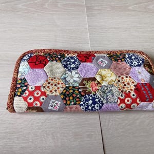 Puede incluir: Un estuche rectangular de patchwork con una variedad de piezas de tela hexagonales de colores. El estuche presenta un borde rojo con estampado floral y una mezcla de telas florales, de lunares y estampadas en tonos rojos, azules, morados y crema.