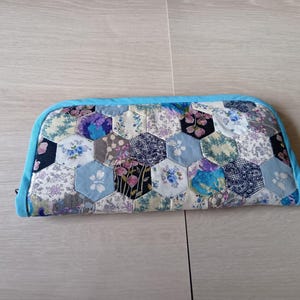 May include: A handmade, quilted clutch with a blue zipper and trim. The clutch is made of hexagonal fabric pieces in various floral patterns, including blue, purple, and white. The overall design is a patchwork style.