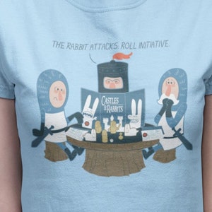 May include: A light blue t-shirt with a graphic of three knights playing a game called "Castles & Rabbits". The text "The rabbit attacks, roll initiative" is printed above the graphic.