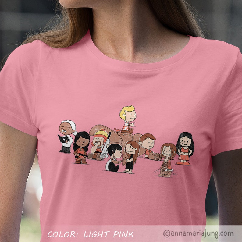 以下が含まれることがあります： ライトピンクのTシャツに、スターウォーズのキャラクターに扮したピーナッツのキャラクターの漫画イラストが描かれています。キャラクターはチャーリー・ブラウン、ルーシー、ライナス、スヌーピー、サリー、ペパーミント・パティ、フランクリンです。シャツの下部に「COLOR: LIGHT PINK」の文字があります。