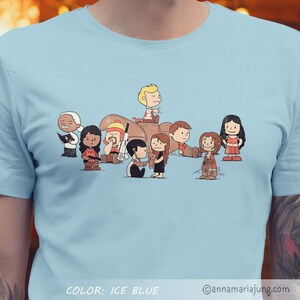 以下が含まれることがあります： ウォーキング・デッドのテレビ番組のキャラクターに扮したピーナッツのキャラクターの漫画のイラストが描かれた薄い青色のTシャツ。Tシャツの下部に「COLOR: ICE BLUE」の文字があります。