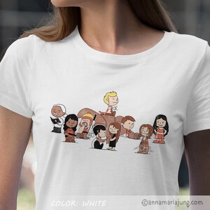 以下が含まれることがあります： ゴーストバスターズに扮したピーナッツのキャラクターの漫画イラストが描かれた白いTシャツ。キャラクターはベージュのジャンプスーツを着て、プロトンパックを持っています。Tシャツの下部に「COLOR: WHITE」の文字があります。