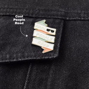 May include: A metal enamel pin shaped like a folded ribbon with three horizontal segments. The pin features the text "COOL PEOPLE READ" in gold lettering, with a small pair of black sunglasses icon next to the word "COOL". The ribbon segments are colored in pale green, peach, and white.
