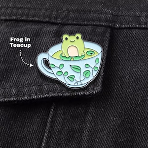 May include: A metal enamel pin featuring a light green frog sitting inside a pale blue teacup filled with green liquid and lily pads. The exterior of the teacup is decorated with a delicate green vine pattern. The text Frog in Teacup is visible.