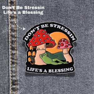 May include: A metal enamel pin featuring a green frog lounging in front of a large orange sun and two mushrooms with red and pink caps. The black border contains the white text "DON'T BE STRESSIN" at the top and "LIFE'S A BLESSING" at the bottom.