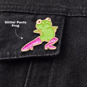 May include: A gold-rimmed enamel pin featuring a light green frog with a rounded belly and arms on hips. The frog wears bright pink glitter trousers that extend down to the feet. The text Glitter Pants Frog is printed to the left of the pin.