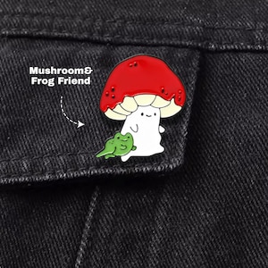 May include: A metal enamel pin featuring a white mushroom character with a large red cap and a small green frog friend. The pin includes the text Mushroom & Frog Friend. The design has black outlines and a smooth finish, attached to black fabric.
