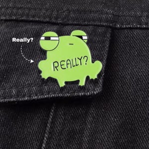 Könnte beinhalten: Eine hellgrüne Emaille-Anstecknadel in Form eines Frosches mit skeptischem Gesichtsausdruck. Der Frosch hat große Augen mit schwarzen Pupillen und den Text "REALLY?" in Schwarz auf seinem Körper. Die Anstecknadel ist an einem schwarzen Stoff befestigt.