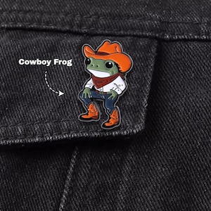 May include: An enamel pin featuring a green frog wearing an orange cowboy hat, a red bandana, a white shirt, blue jeans, and orange cowboy boots. The pin is attached to black denim fabric. The text Cowboy Frog is printed to the left of the pin.
