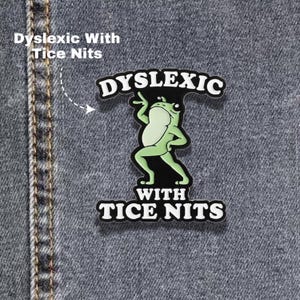 May include: A black and light green enamel pin featuring a cartoon frog in a dancing pose. The text DYSLEXIC is arched above the frog, and the text WITH TICE NITS is positioned below the frog in bold white capital letters with a black outline.