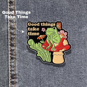 May include: An enamel pin featuring a green frog sitting on a red and beige mushroom, surrounded by flowers and a butterfly. The text "Good things take time" is printed in beige on a black background. The pin is attached to dark blue denim fabric.